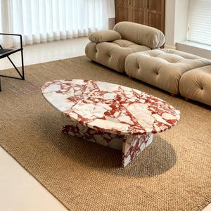 Op de afbeelding: Ovale marmeren salontafel met een rood en wit patroon, geplaatst op een geweven jute vloerkleed. Een zwarte metalen stoel en een beige hoekbank staan op de achtergrond, wat een moderne woonkamer creëert.