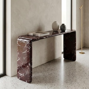 Op de afbeelding: Een moderne console met een bordeauxrode marmeren afwerking en witte adering. Twee vazen en een boek staan op het tafelblad. Een gouden vloerlamp staat in de buurt. De tafel is ongeveer 122 cm lang.