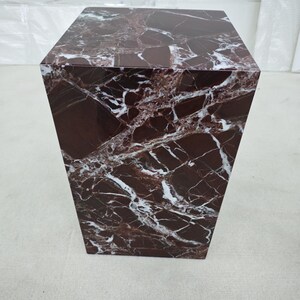 May include: A square, marble-like pedestal table with a rich, dark burgundy color and striking white veining. The table has a solid, geometric design, suitable for displaying decorative items or as a side table. The overall aesthetic is modern and luxurious.