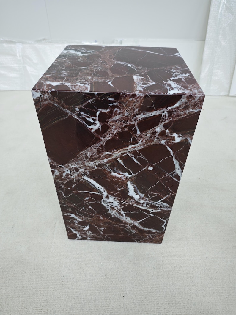 May include: A square, marble-like pedestal table with a rich, dark burgundy color and striking white veining. The table has a solid, geometric design, suitable for displaying decorative items or as a side table. The overall aesthetic is modern and luxurious.