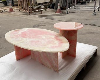 May include: A set of two pink onyx stone tables. The larger table is oval-shaped, and the smaller table is round. Both tables have a pink and white marbled pattern and are supported by rectangular bases.