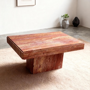 Puede incluir: Una mesa de centro cuadrada, de color marrón rojizo, con una base sólida en forma de bloque. La mesa tiene una parte superior rectangular con un patrón de vetas de madera visible. La mesa está colocada sobre una alfombra beige claro, con un cuadro enmarcado y objetos decorativos en el fondo.