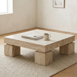 Pode incluir: Uma mesa de centro quadrada de pedra clara, com um livro e uma caneca em cima. A mesa tem uma base em bloco e assenta num tapete de tons neutros. Uma pequena imagem emoldurada está pendurada na parede.