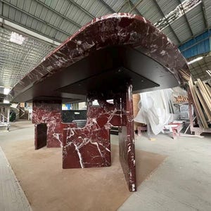 May include: A large, modern table with a dark red marble top and matching supports. The marble has white veining, and the table is set in a warehouse environment. The table's dimensions are not visible.