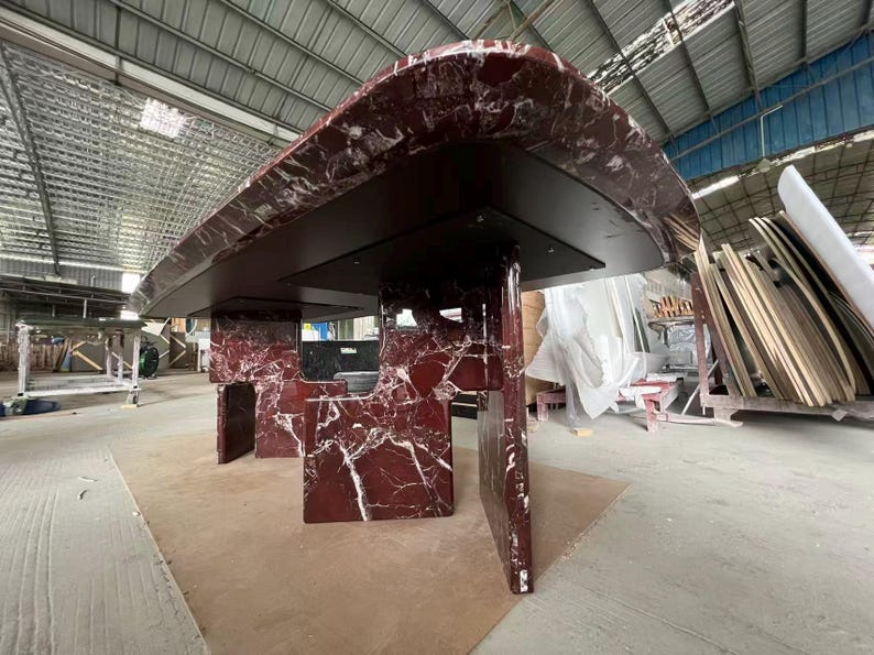 May include: A large, modern table with a dark red marble top and matching supports. The marble has white veining, and the table is set in a warehouse environment. The table's dimensions are not visible.