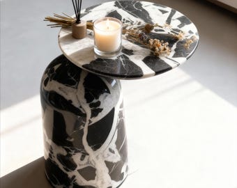 May include: A modern side table with a cylindrical base and a round top, both featuring a black and white marble pattern. A lit candle, dried flowers, and a reed diffuser sit atop the table, creating a minimalist aesthetic.