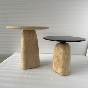 May include: Two stone-like side tables. One has a light beige, round top and a matching base. The other has a black, round top and a beige base. The bases are textured and have a rounded, organic shape. The tables are likely made of stone or a stone-like material.