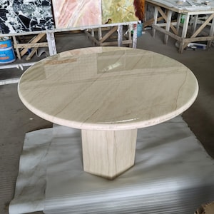 May include: A round table with a polished beige stone top featuring subtle linear veining and a smooth, rounded edge. The table is supported by a single, thick, hexagonal pedestal base made of the same light-colored stone material.