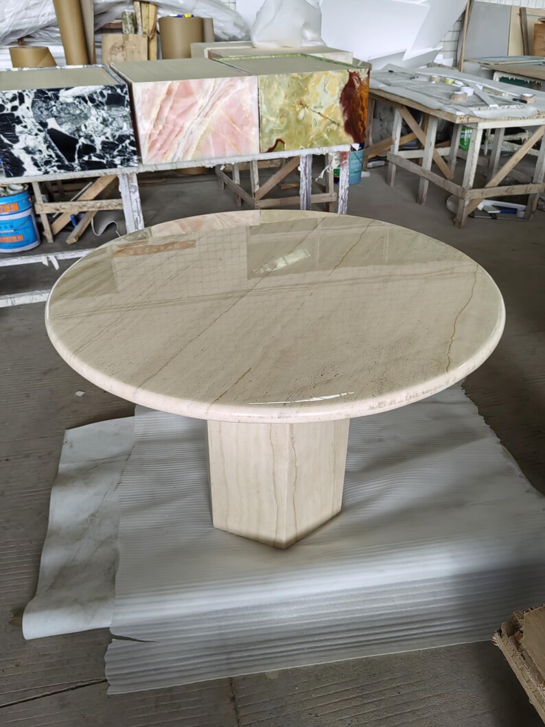May include: A round table with a polished beige stone top featuring subtle linear veining and a smooth, rounded edge. The table is supported by a single, thick, hexagonal pedestal base made of the same light-colored stone material.