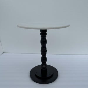 Puede incluir: Pequeña mesa auxiliar redonda con tapa de mármol blanco. La mesa tiene un pedestal de madera torneada negra y una base circular. La mesa mide aproximadamente 61 cm de alto y la tapa mide 46 cm de diámetro.