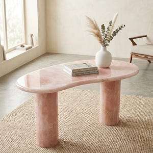 Peut inclure: Une table basse en forme de rein avec une surface rose polie et deux pieds cylindriques. Un vase blanc contenant de l'herbe de la pampa séchée et deux livres empilés intitulés Modernist Stone et WABI-SABI HOME reposent sur la table.