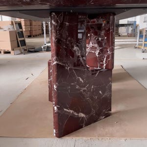 May include: A modern dining table with a dark red marble base and a black tabletop. The base features a unique geometric design with white veining throughout the marble. The table is set in a warehouse setting.
