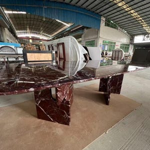 May include: A large, oval-shaped conference table with a polished burgundy marble top and matching geometric base. The table is in a warehouse setting, showcasing its size and design.