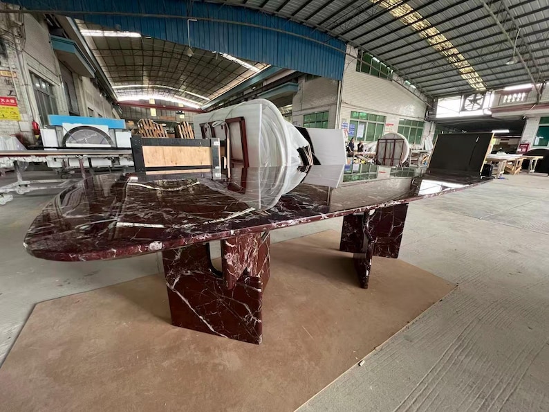May include: A large, oval-shaped conference table with a polished burgundy marble top and matching geometric base. The table is in a warehouse setting, showcasing its size and design.