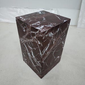 May include: A square, dark red marble pedestal with white veining. The pedestal has a geometric, tapered design, wider at the top and narrower at the base. The surface has a polished finish, showcasing the natural stone's patterns.