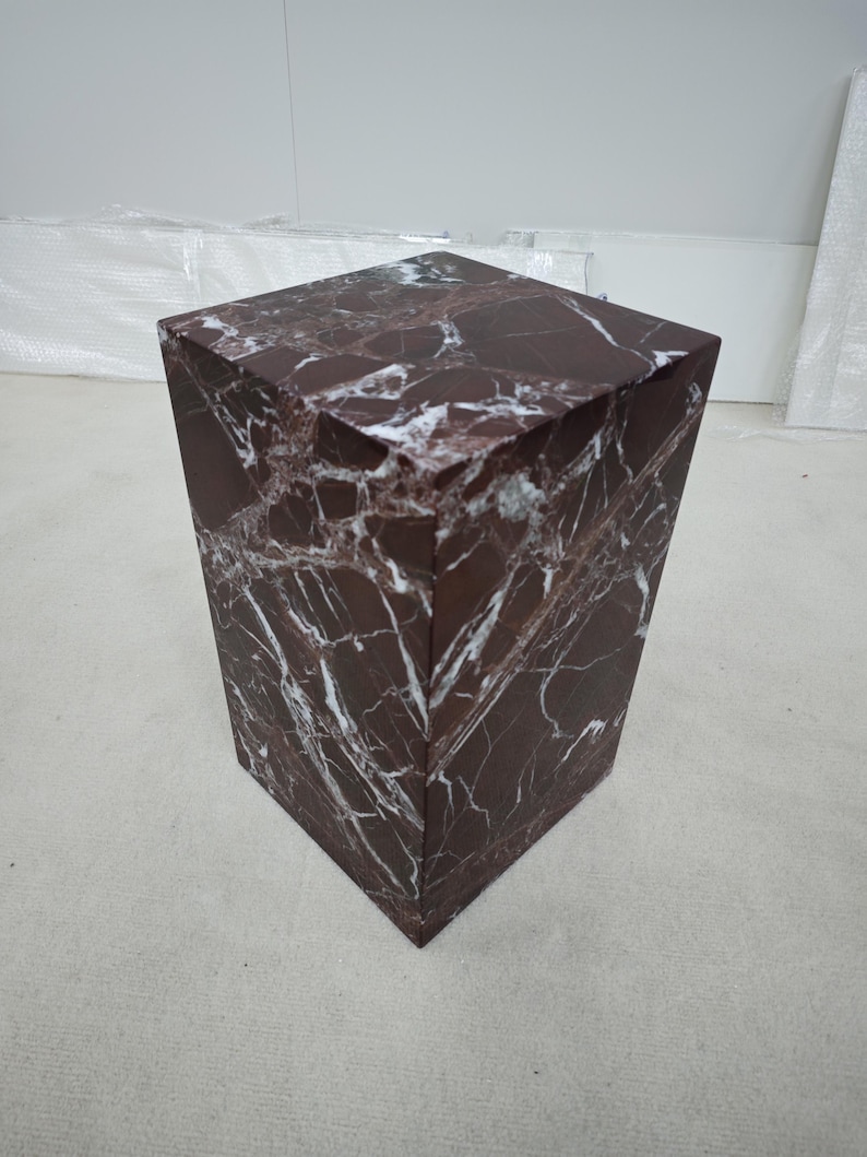 May include: A dark red marble pedestal with white veining. The square-shaped pedestal has a slightly tapered design, wider at the top and narrower at the base. The marble has a polished finish, reflecting light.