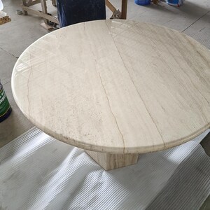 May include: A round beige travertine stone table with a polished surface featuring natural veining and a smooth, rounded edge. The table is supported by a central pedestal base made of the same light-colored, textured stone material.