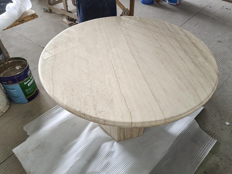May include: A round beige travertine stone table with a polished surface featuring natural veining and a smooth, rounded edge. The table is supported by a central pedestal base made of the same light-colored, textured stone material.