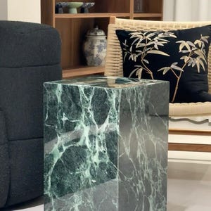 Puede incluir: Una mesa auxiliar cuadrada de mármol verde oscuro con vetas blancas. La mesa tiene una superficie pulida y está colocada en un salón. La mesa es una pieza decorativa.