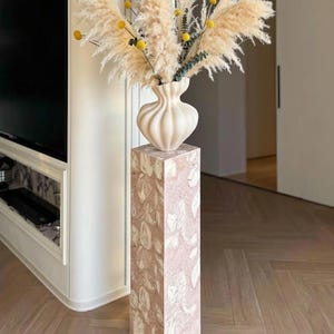 Puede incluir: Un arreglo decorativo con un jarrón crema con diseño acanalado, que contiene hierba de la pampa seca, botones de billy amarillos y eucalipto. El jarrón se encuentra sobre un pedestal rectangular alto con un patrón de mármol rosa y blanco.