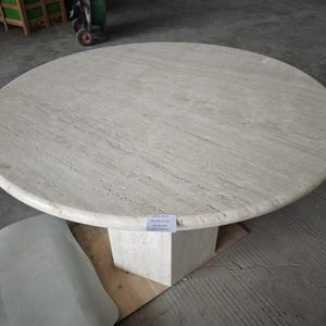 May include: A round, beige travertine stone table with a natural porous texture and visible veining. The tabletop is supported by a thick, square-shaped pedestal base made of the same light-colored stone material, featuring a small white label.