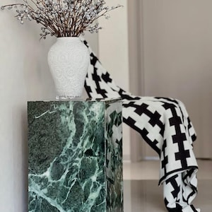 Puede incluir: Arreglo decorativo con un pedestal de mármol verde, un jarrón blanco con ramas con pequeños acentos plateados. Una manta estampada en blanco y negro está drapeada sobre el pedestal.