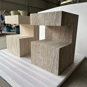 May include: A set of three-tiered, geometric display stands. The stands are constructed from a light brown, textured material, possibly stone or concrete, with a natural, earthy appearance. The design features a stepped arrangement, suitable for showcasing products.