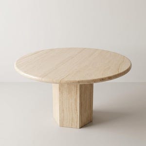 May include: A round coffee table crafted from beige travertine stone. The table features a smooth, circular tabletop with visible natural veining and a thick, hexagonal pedestal base that provides structural support for the surface above.