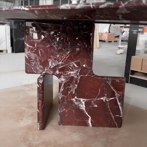 May include: A modern table with a rich burgundy marble top and matching base. The base features a unique geometric design with a rectangular support and a cutout section. White veining adds contrast to the dark red surface.
