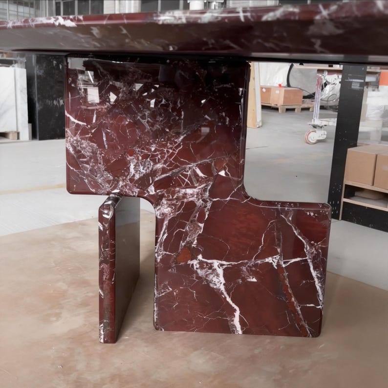 May include: A modern table with a rich burgundy marble top and matching base. The base features a unique geometric design with a rectangular support and a cutout section. White veining adds contrast to the dark red surface.