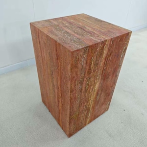 May include: A solid, reddish-brown wooden cube-shaped stool with a natural grain pattern. The stool has a slightly trapezoidal shape, with one side angled inward. The wood's color varies from light to dark brown.