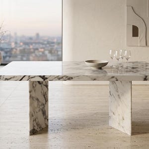 May include: A rectangular marble dining table with a grey and white veined pattern. The table has a thick top and two pedestal legs. A bowl and wine glasses are on the table. The room has a large window with a city view.