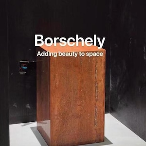 May include: A reddish-brown stone block with the text "Borschely" and "Adding beauty to space" in white. The block is rectangular and stands on a white surface, set against a dark background.