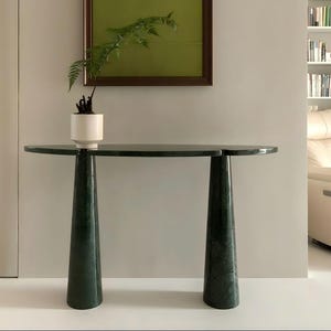 Op de afbeelding: Een donkergroene marmeren console met een halfronde bovenkant en twee conische poten. Een witte keramische pot met een groene plant staat op de tafel. Een groen kunstwerk met lijst hangt aan de muur achter de tafel.