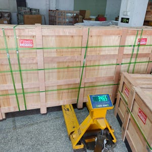 May include: Large wooden shipping crates secured with green straps and labeled with "Fragile" stickers. A yellow pallet jack with a digital scale displaying a weight of 3465 is positioned beneath the crates, indicating a warehouse or shipping environment.