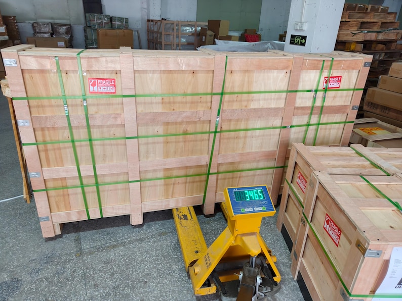 May include: Large wooden shipping crates secured with green straps and labeled with "Fragile" stickers. A yellow pallet jack with a digital scale displaying a weight of 3465 is positioned beneath the crates, indicating a warehouse or shipping environment.