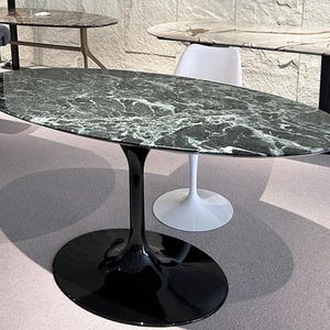 May include: An oval-shaped dining table with a black glossy base and a green and white marble top. The table is surrounded by other tables with similar designs, showcasing a modern furniture collection.