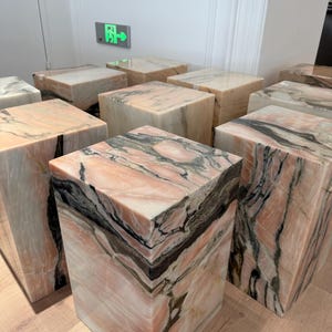 Puede incluir: Colección de taburetes cúbicos de mármol. Los taburetes presentan una mezcla de vetas rosas, blancas y negras. La piedra natural tiene un acabado pulido y los cubos están dispuestos sobre un suelo de madera clara.