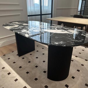 May include: An oval dining table with a black and white marble top. The table has two black, solid, rectangular legs and a black, cylindrical, ribbed leg. The table sits on a light-coloured rug with dark brown square patterns.