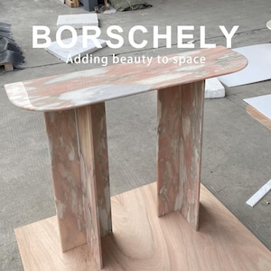 May include: A pink and white marble console table with a semi-circular top and two vertical supports. The table has the text "BORSCHELY" and "Adding beauty to space" in white letters.