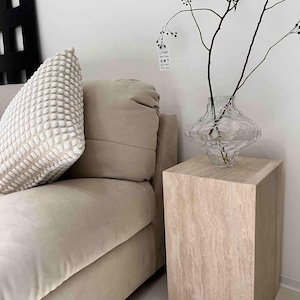 May include: A beige sofa with a textured pillow and a decorative glass vase holding branches. The vase sits on a light-colored, square side table. The overall aesthetic is minimalist and modern.
