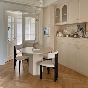 May include: A round dining table with a textured beige pedestal base and three chairs featuring dark wood frames and cream-colored upholstered seats. A white spherical pendant light hangs above, and a beige cabinet unit stands against the wall.