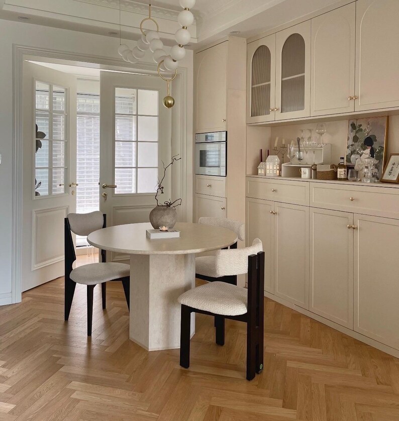 May include: A round dining table with a textured beige pedestal base and three chairs featuring dark wood frames and cream-colored upholstered seats. A white spherical pendant light hangs above, and a beige cabinet unit stands against the wall.