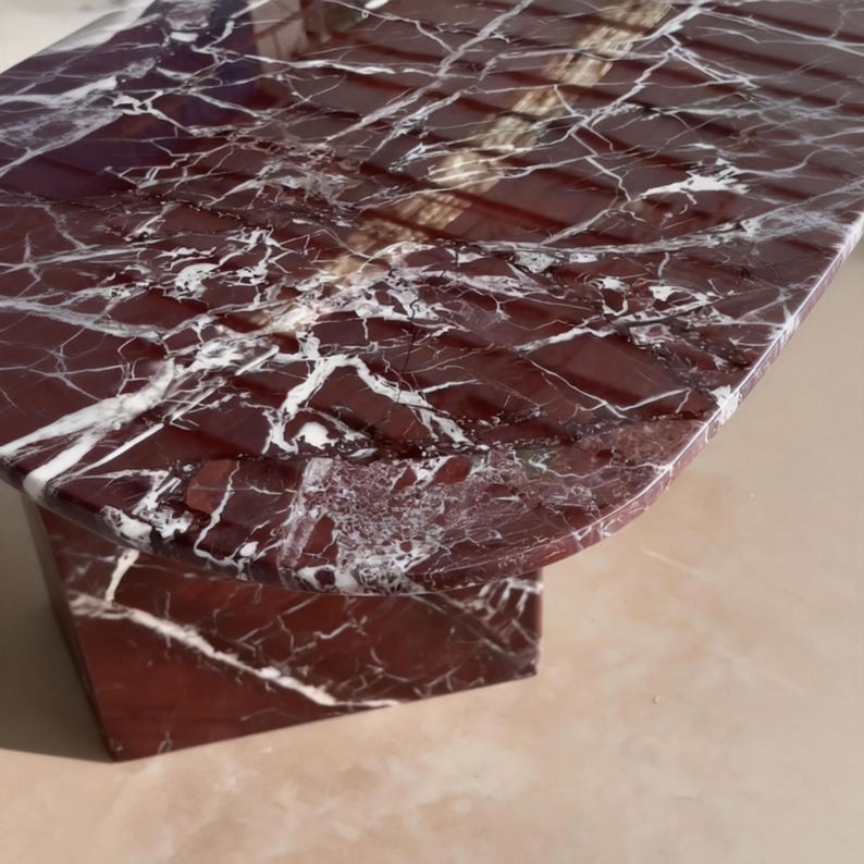 May include: A close-up of a red marble table with white veining. The table has a rectangular top with rounded edges and a solid base. The polished surface reflects light, highlighting the natural patterns.