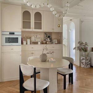 May include: A round stone dining table with a central pedestal base, surrounded by three chairs featuring black wooden frames and white textured upholstery. Above the table hangs a long, curved light fixture with white spherical glass globes.