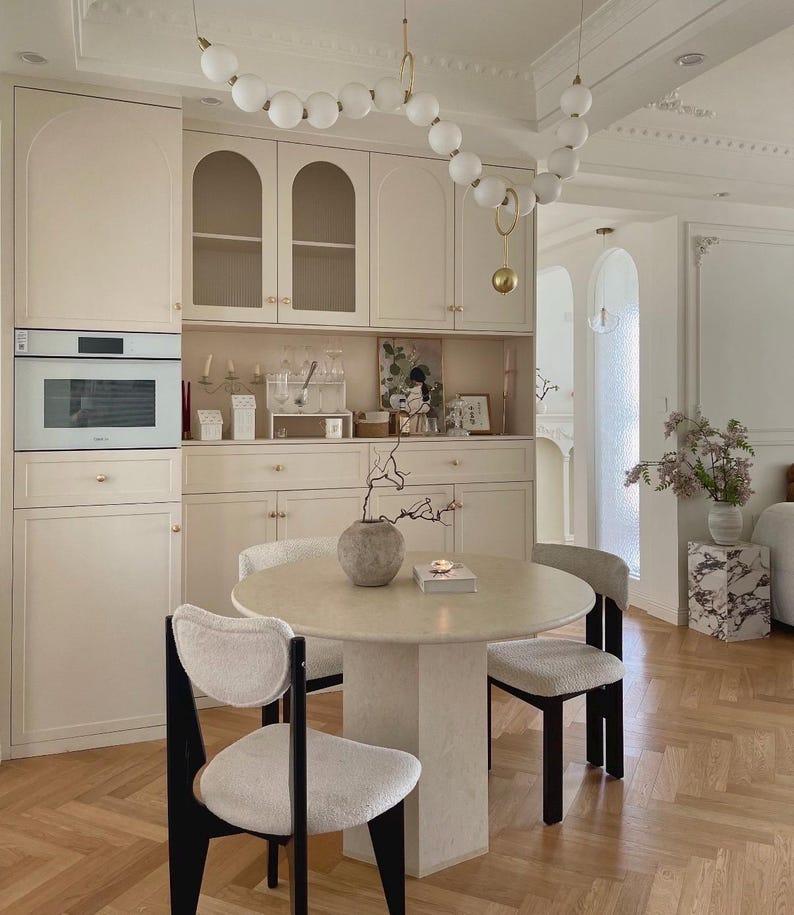 May include: A round stone dining table with a central pedestal base, surrounded by three chairs featuring black wooden frames and white textured upholstery. Above the table hangs a long, curved light fixture with white spherical glass globes.