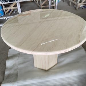 May include: A round table with a polished beige stone top featuring subtle brown veining. The table is supported by a single, thick, hexagonal pedestal base made of the same beige stone, creating a cohesive and solid structure for the furniture piece.