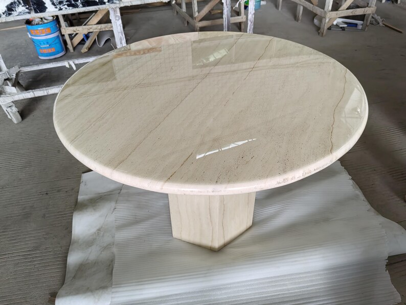 May include: A round table with a polished beige stone top featuring subtle brown veining. The table is supported by a single, thick, hexagonal pedestal base made of the same beige stone, creating a cohesive and solid structure for the furniture piece.
