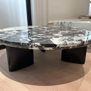 May include: A modern, low coffee table with a round, marble top featuring black and white veining. The top rests on three black, curved legs. The table is set on a light wood floor.