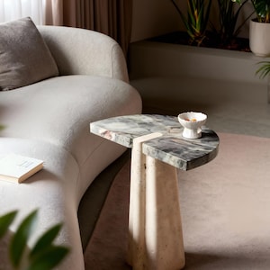Puede incluir: Una mesa auxiliar moderna con una tapa de mármol en tonos grises, blancos y verdes. La mesa tiene una base geométrica única hecha de piedra de color claro. Un pequeño cuenco blanco está sobre la mesa, con un sofá curvo en el fondo.